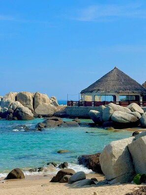 Una playa con rocas grandes, agua turquesa cristalina y un pequeño edificio con techo de paja.