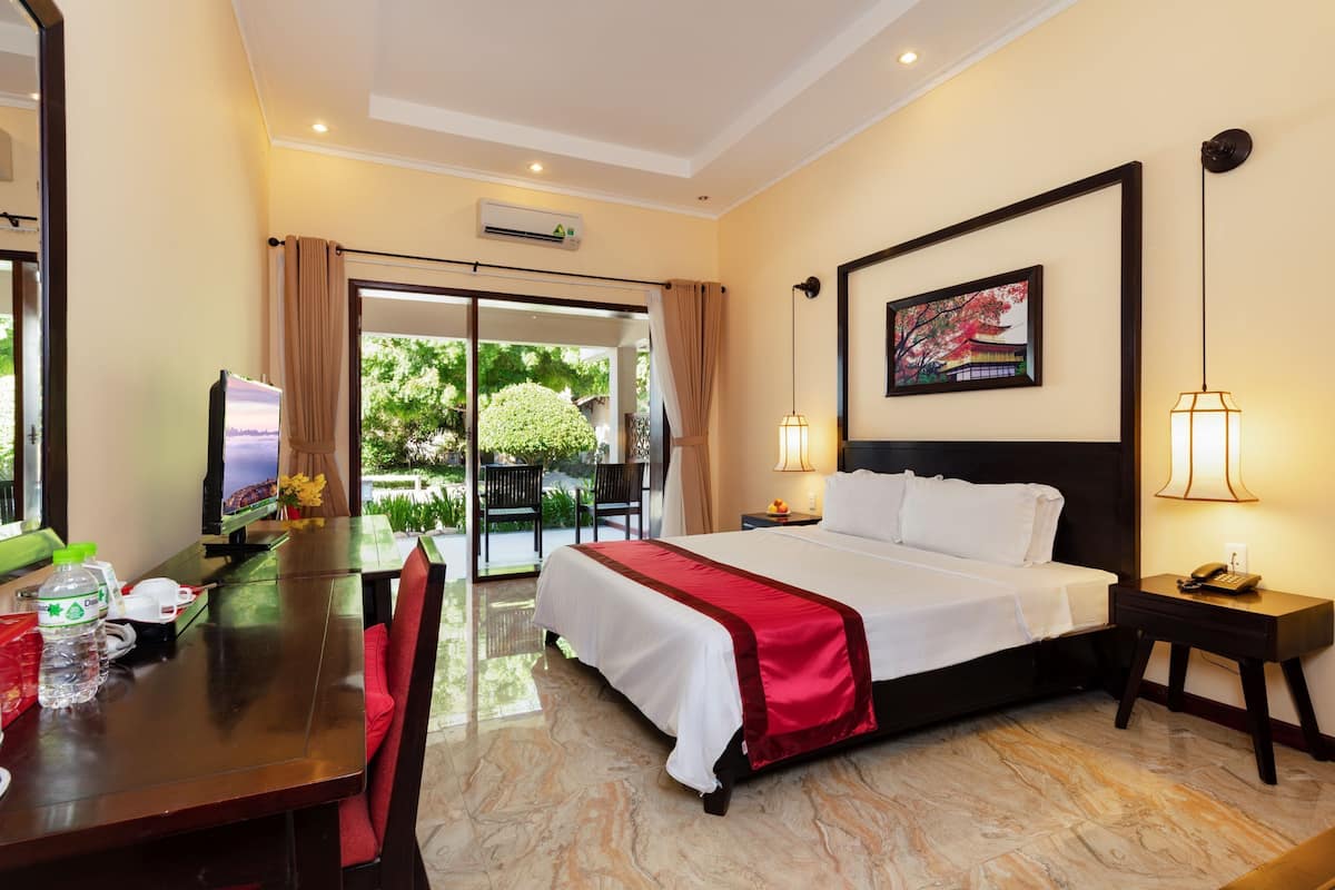 A hotel room with a bed, a desk with a computer, a chair, a table with a bottle, and a view of greenery outside.