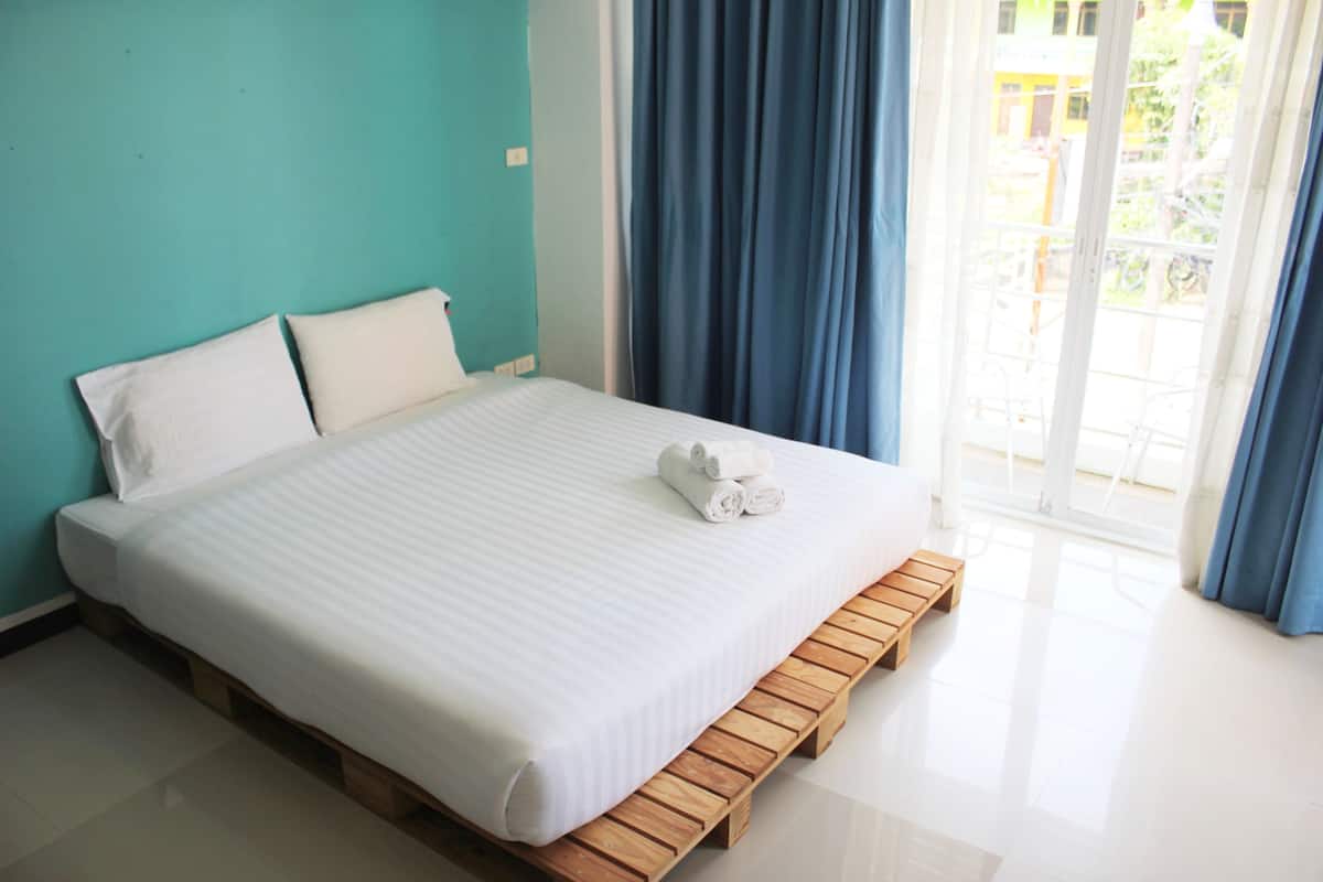 A neatly made bed with a wooden platform, white bedding, and a view of a balcony through a window with blue curtains.
