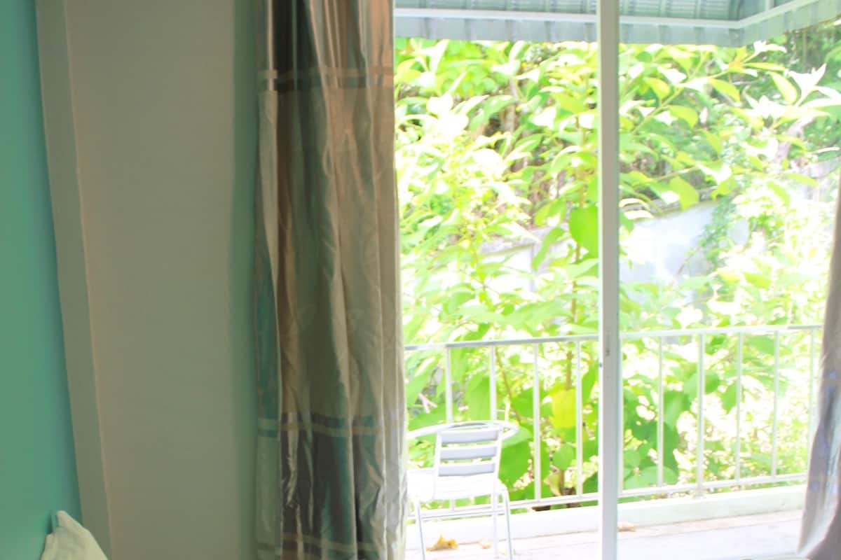 A hotel room with a bed, a window with curtains, and a view of greenery outside.