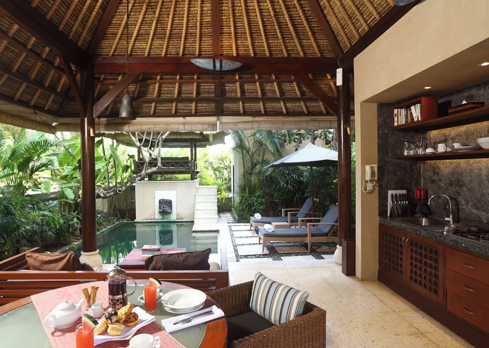 A poolside area with a dining table set for breakfast, lounge chairs, and a kitchen area with a stone backsplash.