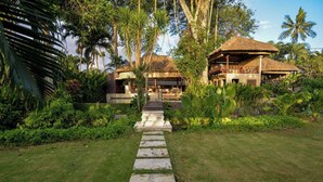 Un resort tropicale con un edificio a tetto di paglia, un vialetto in pietra che conduce all'ingresso e una vegetazione lussureggiante tutt'intorno.