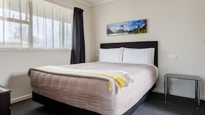 A neatly made bed with a beige quilt, a small bedside table, a painting of a mountain landscape, and a window with sheer curtains.