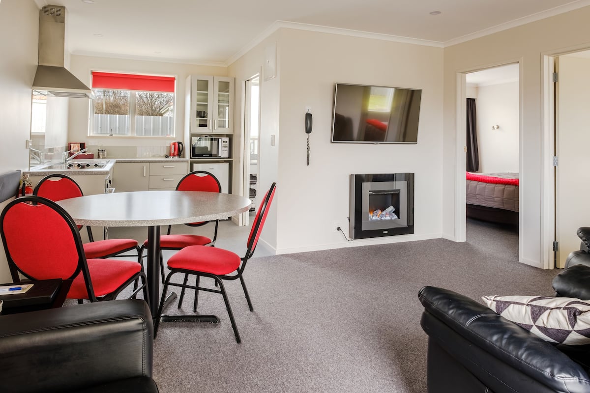 A modern living room with a fireplace, a dining area with red chairs, and a kitchen in the background.