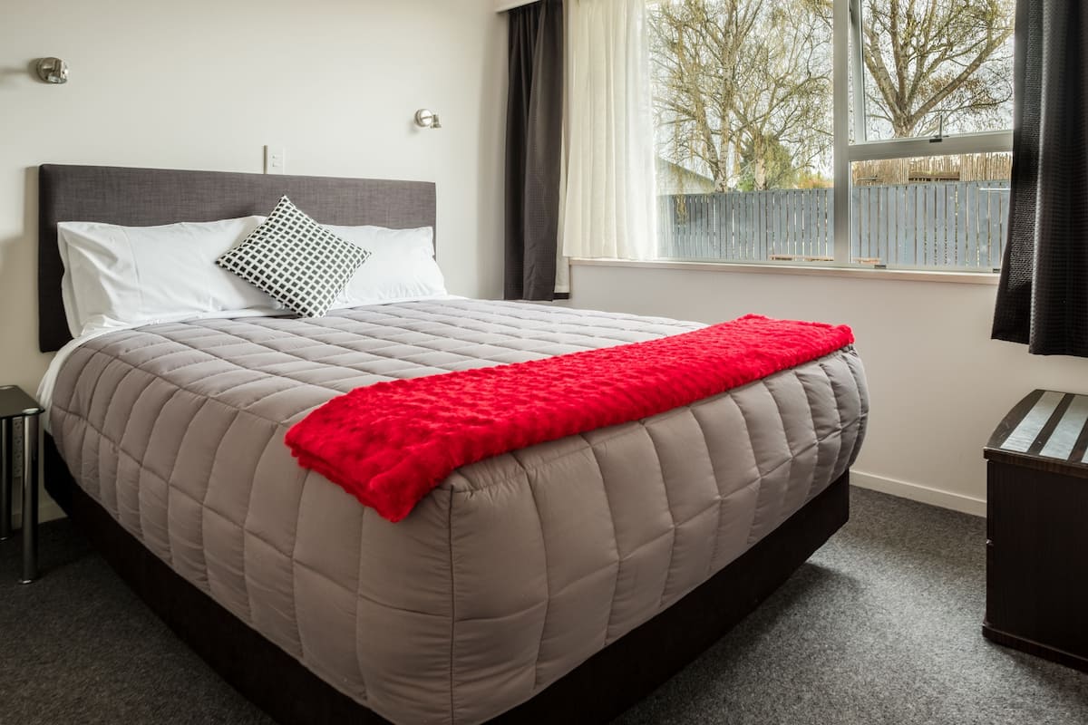 A bed with a quilted headboard, a red throw blanket, and a window with a view of trees and a fence.