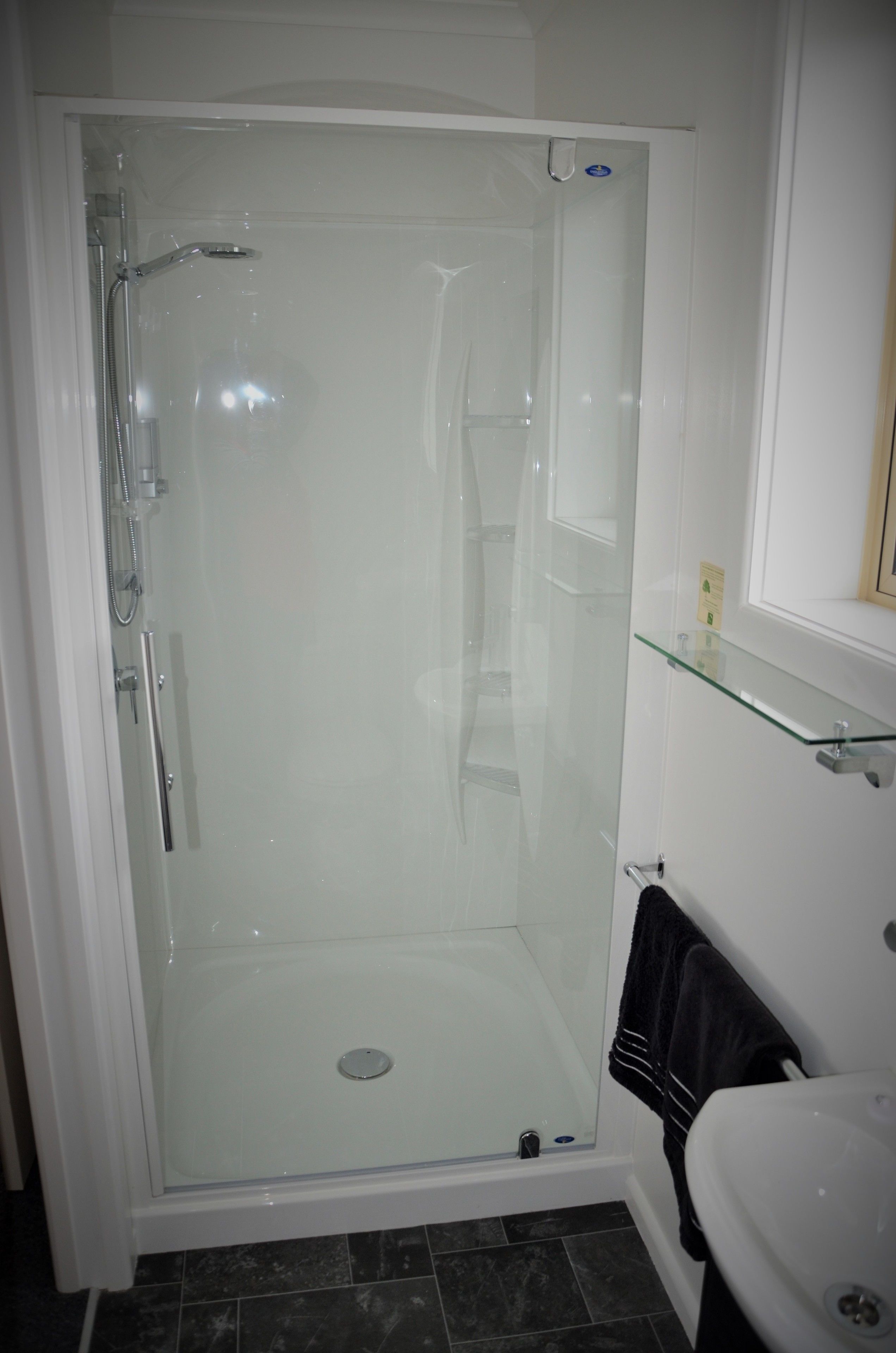 A glass-enclosed shower stall with a handheld showerhead, a glass shelf above the sink, and a black towel hanging on a rack.