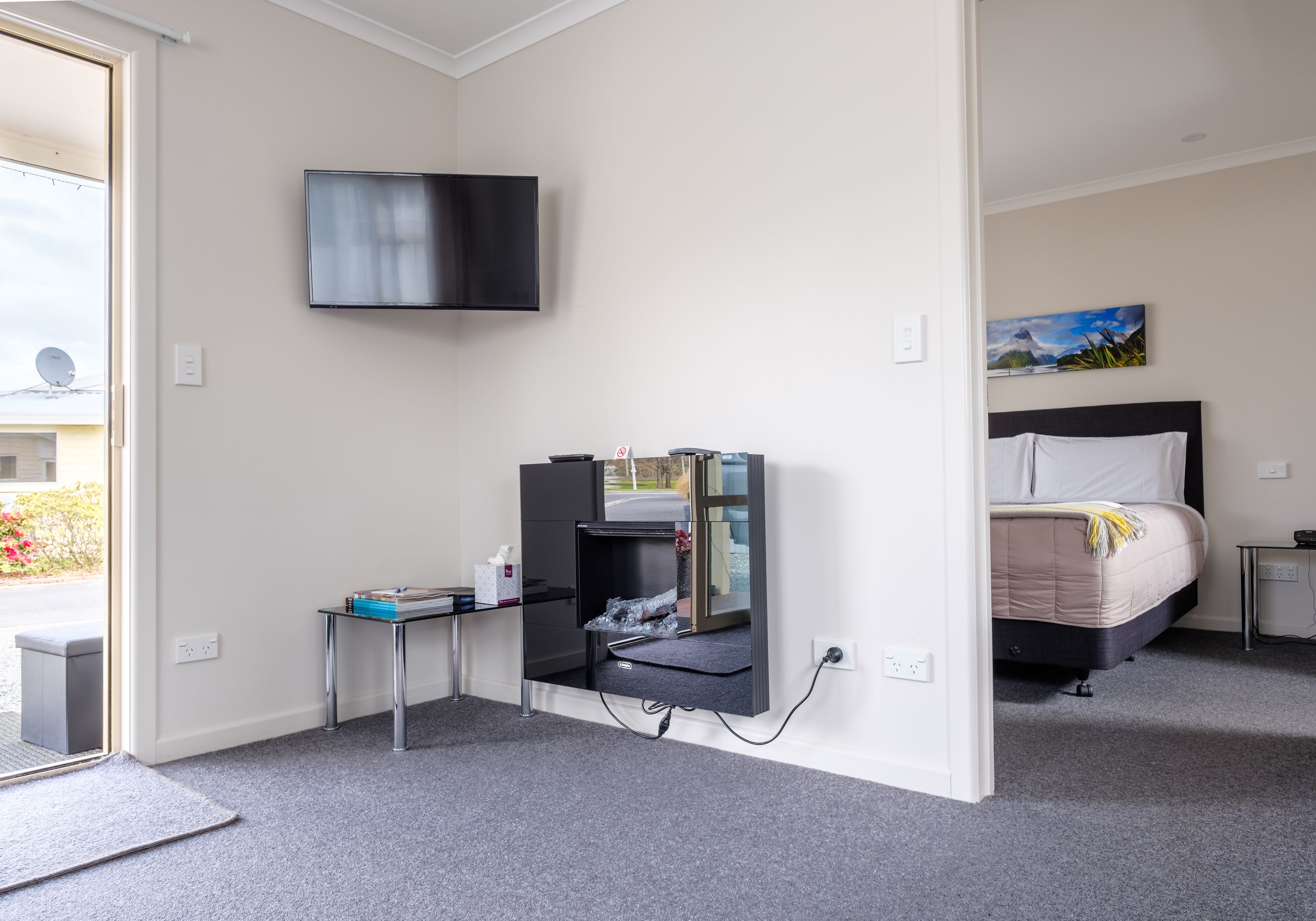 A hotel room with a bed, a TV mounted on the wall, a desk with a computer, and a small table with a chair.