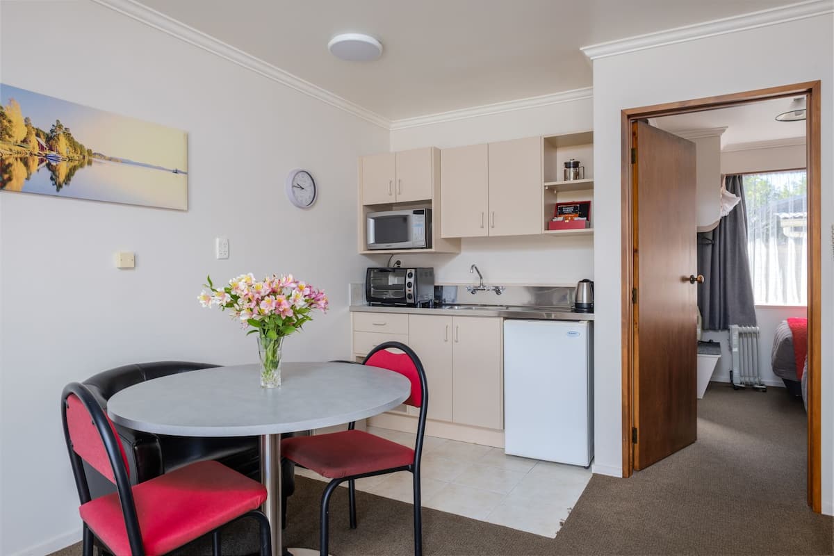 A compact kitchen with a microwave, stove, and refrigerator. There is a dining area with a table and chairs, and a small window with curtains.