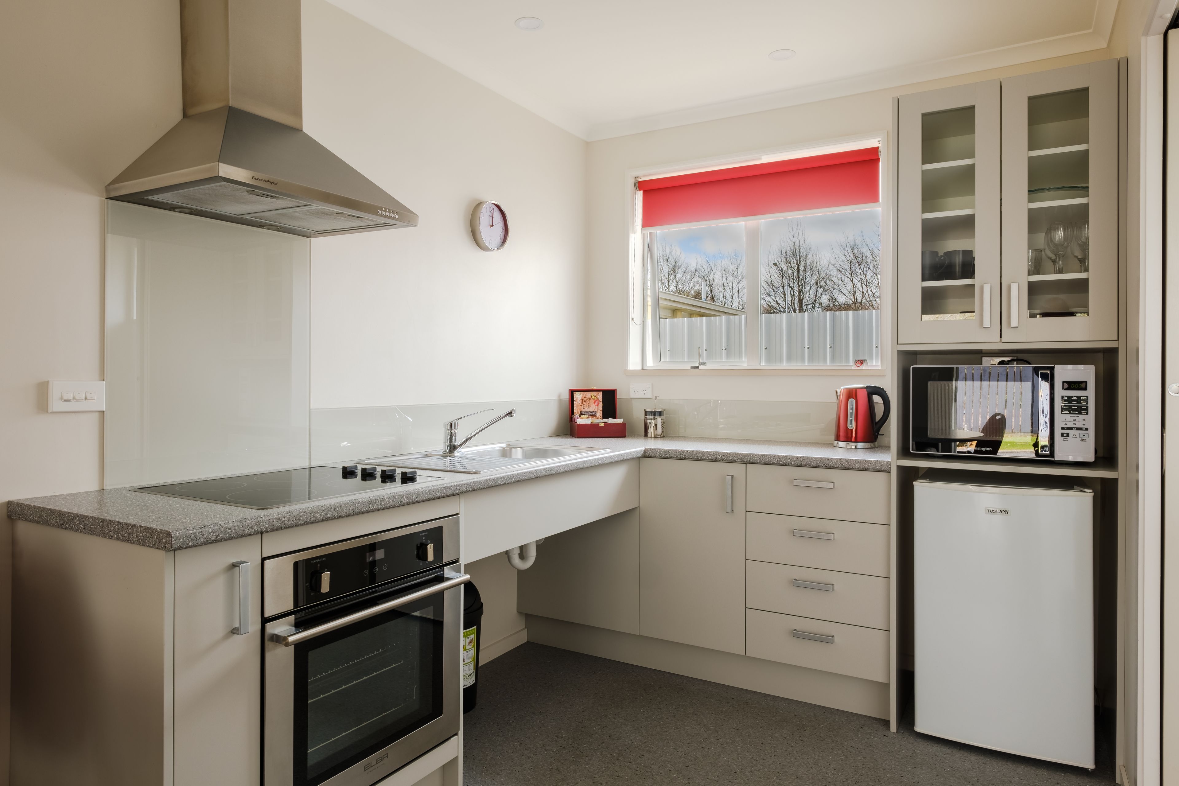 Een moderne keuken met een roestvrijstalen afzuigkap, een magnetron, een inbouwoven en een raam met een rode jaloezie.