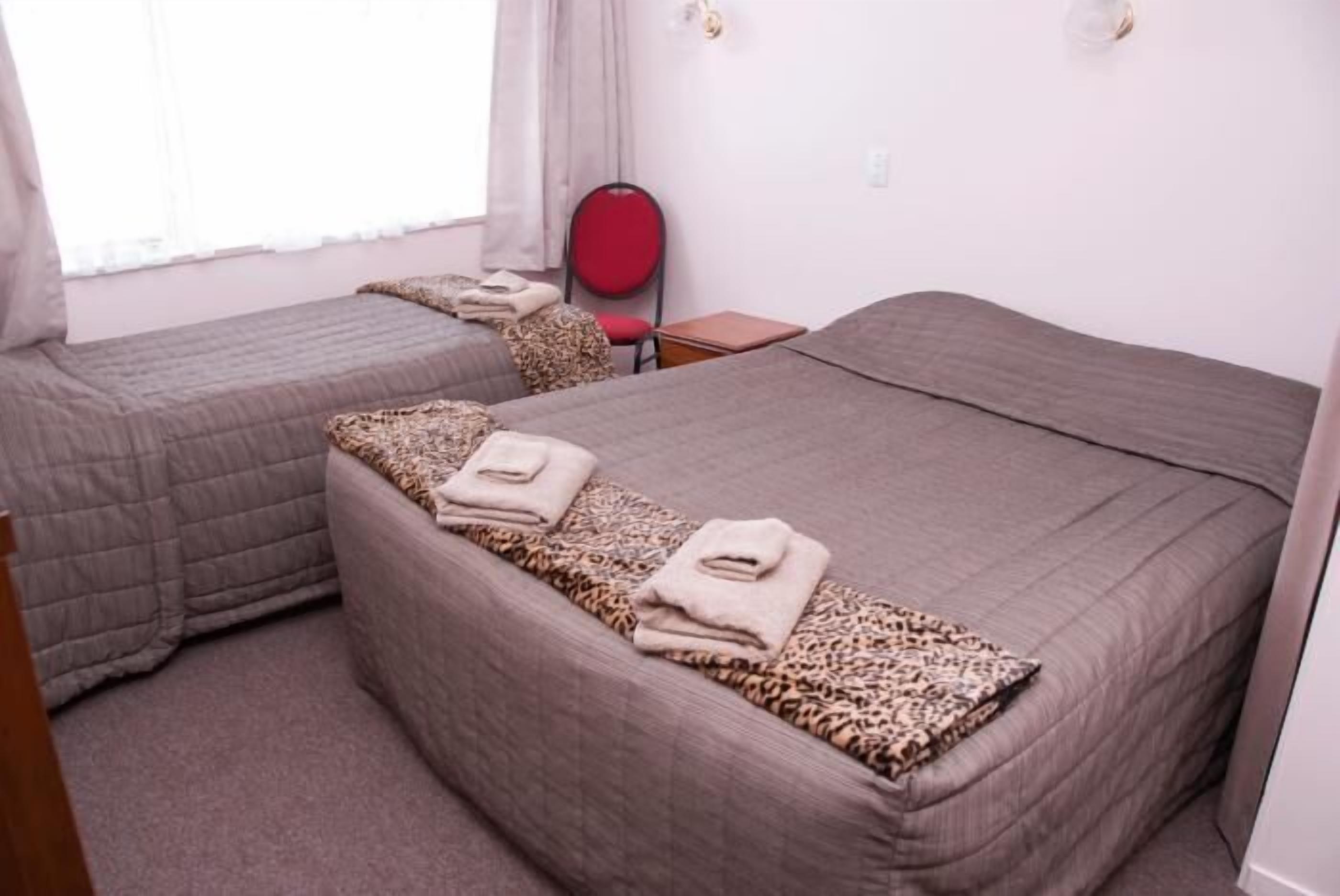 Una camera d'albergo con due letti, ognuno con una coperta a stampa leopardo e un asciugamano piegato. Nell'angolo, un piccolo tavolo e una sedia rossa.