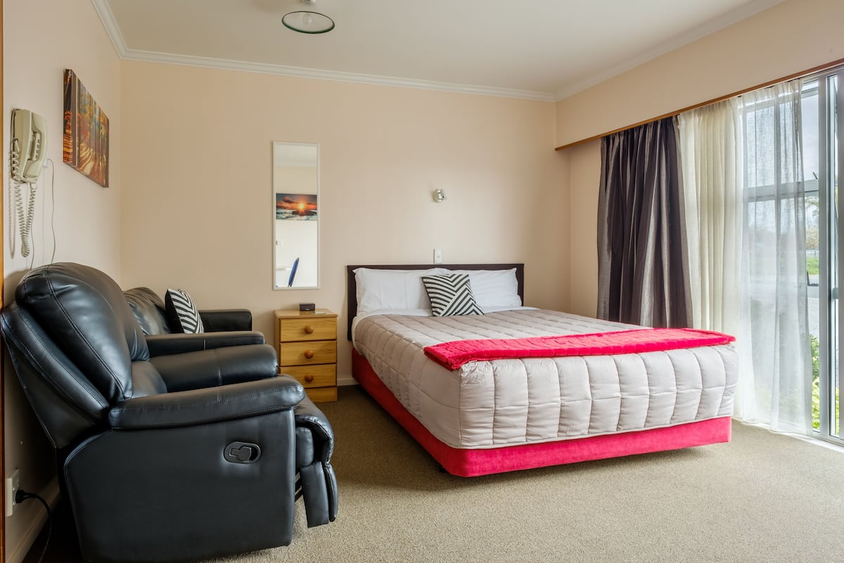 A bedroom with a bed, a black leather chair, a nightstand, a mirror, and a window with curtains.