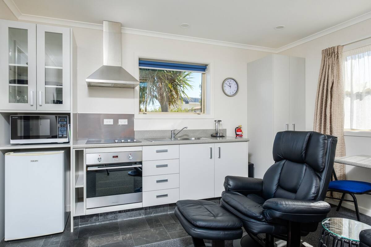 A compact kitchen with white cabinets, a microwave, a built-in oven, a window with a view, and a black leather chair.