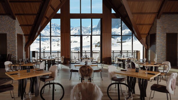 Un restaurant avec des tables et des chaises en bois, de grandes fenêtres donnant sur les montagnes et un mur en pierre.