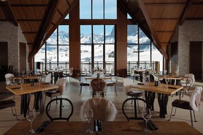 A restaurant with wooden tables and chairs, large windows with a mountain view, and a stone wall.