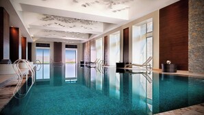 Une piscine intérieure moderne avec des murs en verre transparent, un mur d’accent en bois et un plafond agrémenté d’un luminaire original.
