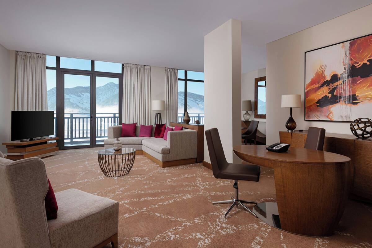 A hotel room with a large window, a sofa, a desk, and a television.