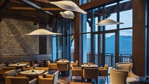 Un espacio interior amplio con varias mesas y sillas, grandes ventanales que ofrecen vistas al exterior, y una estructura de techo de madera.