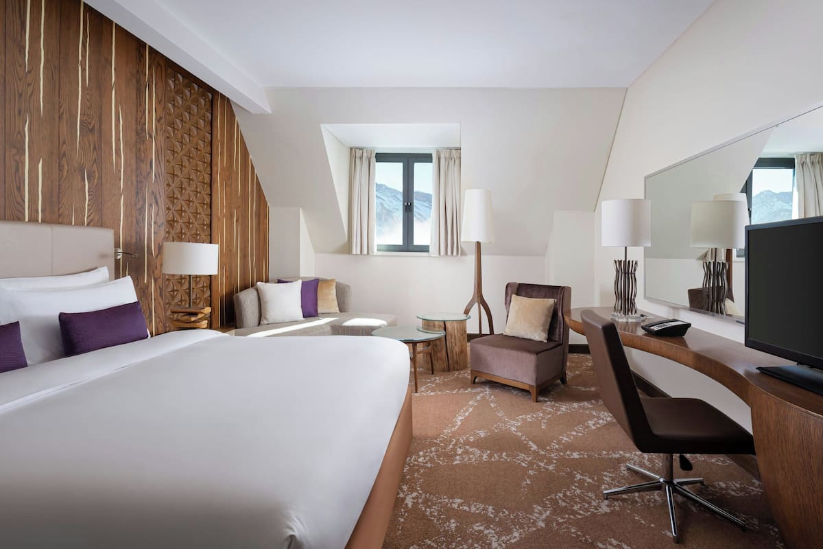 A hotel room with a large bed, a desk with a TV, a chair, a small table, and a window with curtains.