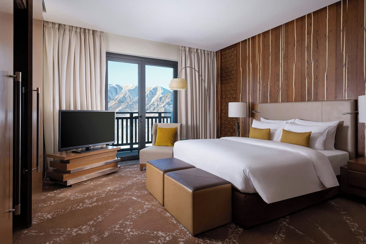 A hotel room with a large bed, a TV, a balcony with a mountain view, and a wooden wall panel.