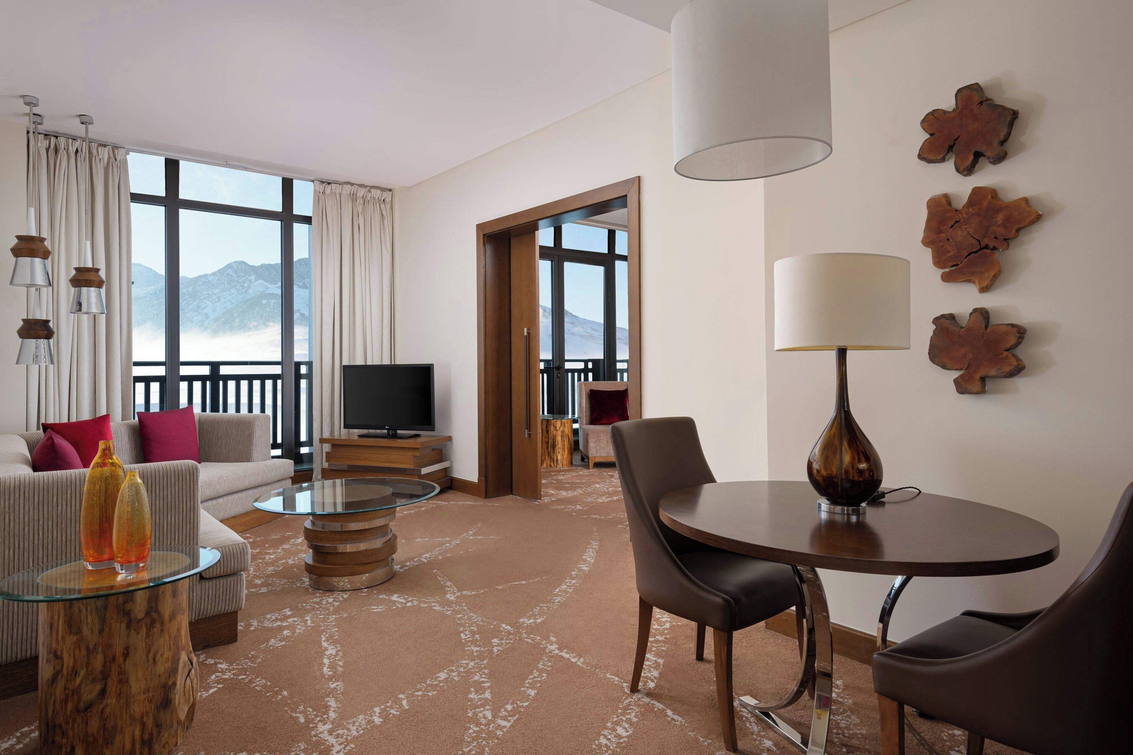 Quarto de hotel moderno com uma grande janela com vista para montanhas, um sofá, uma mesa de centro, uma mesa de jantar com cadeiras e uma televisão.