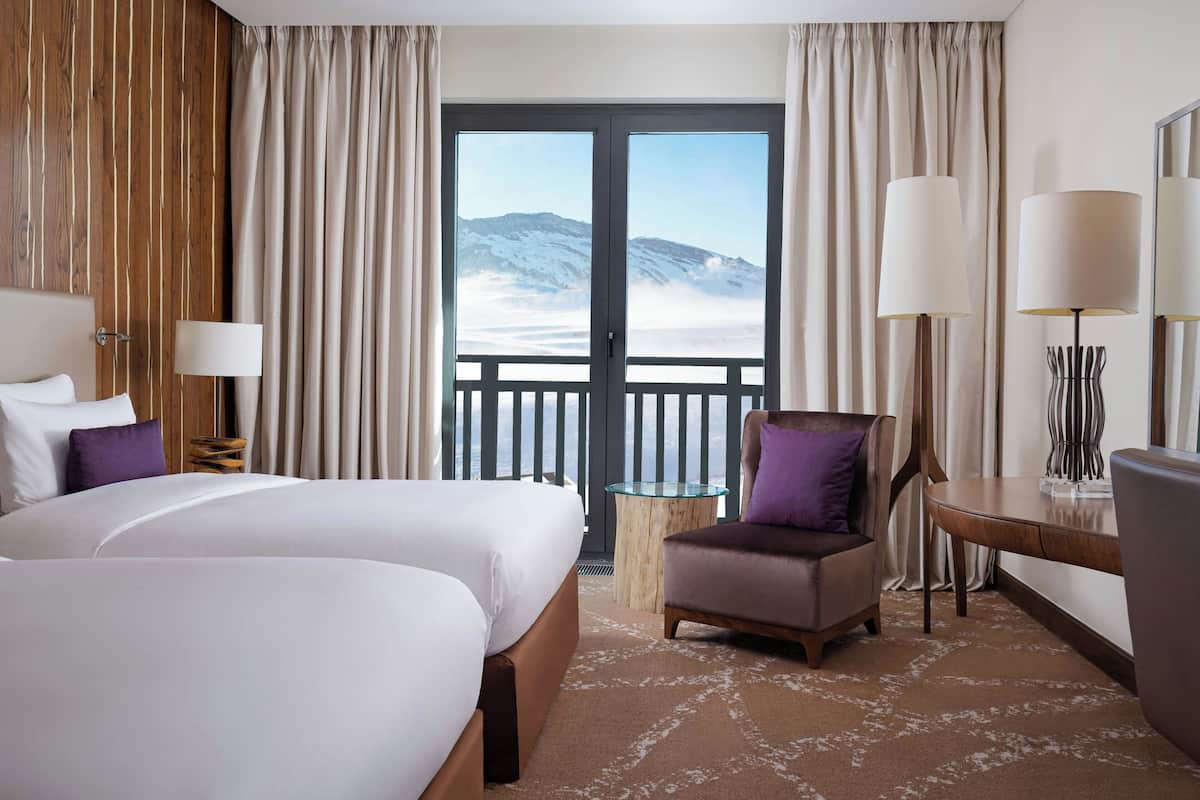 A hotel room with two beds, a chair, a table, and a view of a snowy mountain through a large window.