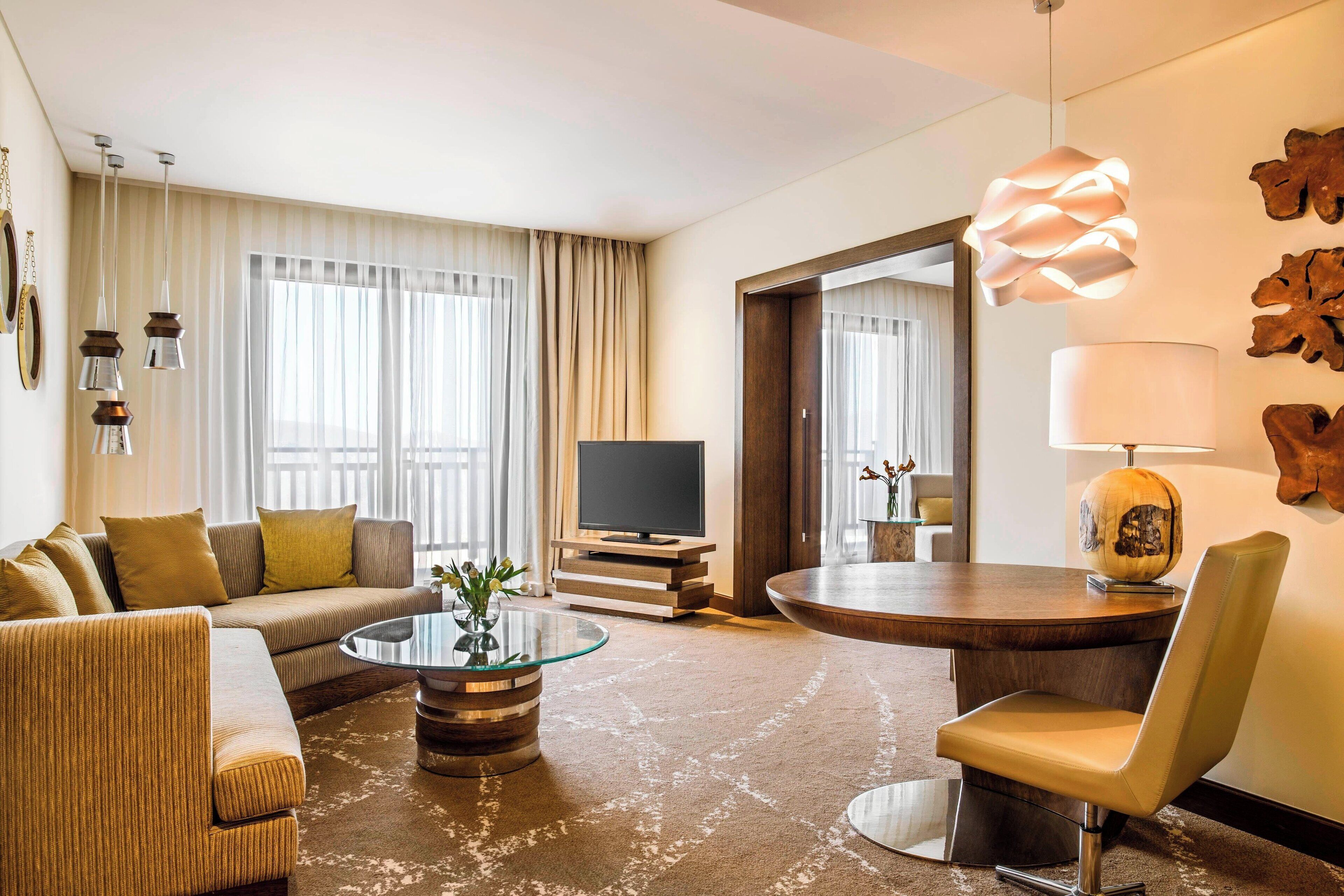 A modern hotel room with a sofa, a glass coffee table, a TV, a dining table with a chair, and a large mirror.
