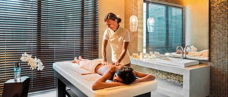 A spa room with a massage table, a person receiving a massage, a large mirror, and a modern sink.
