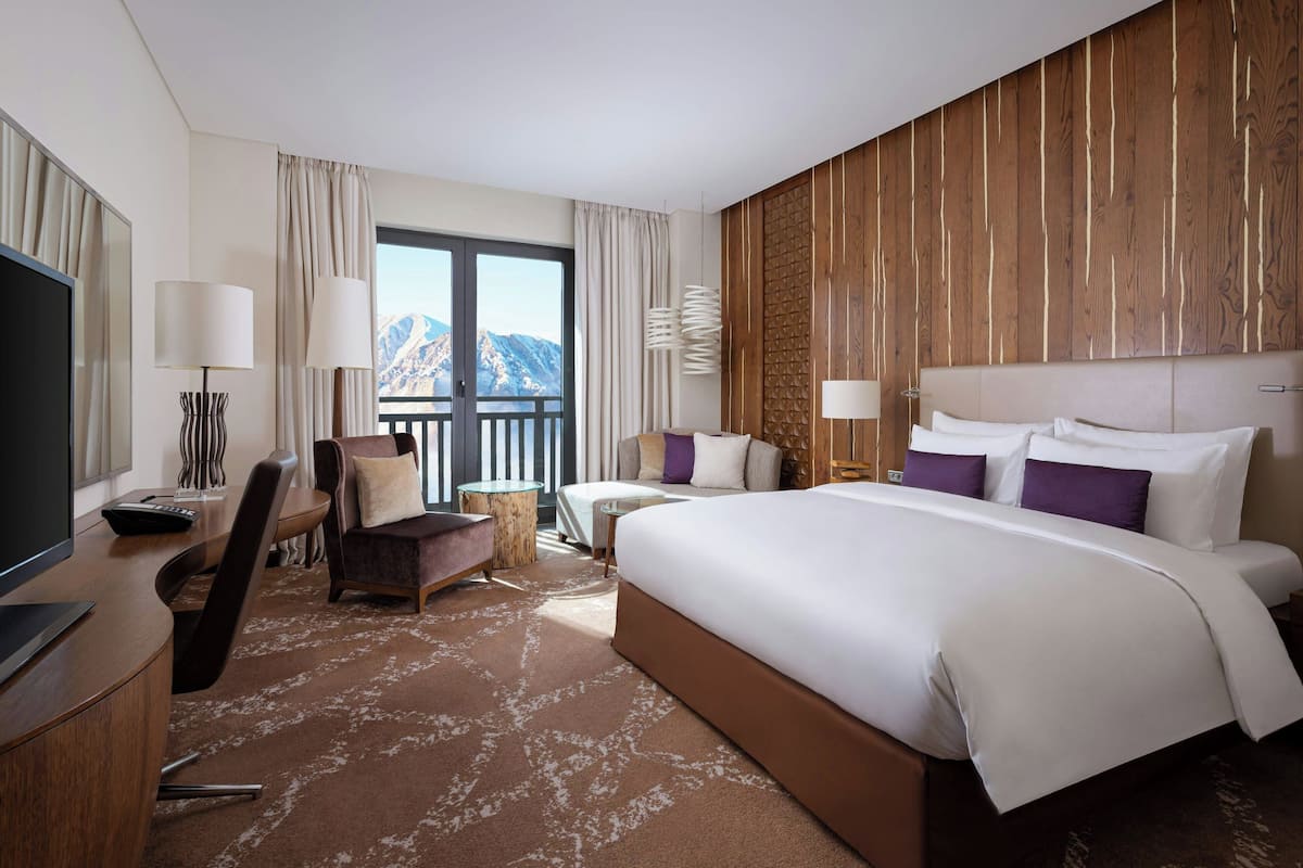 A hotel room with a large bed, a desk with a TV, a chair, a small table, and a view of mountains through the window.