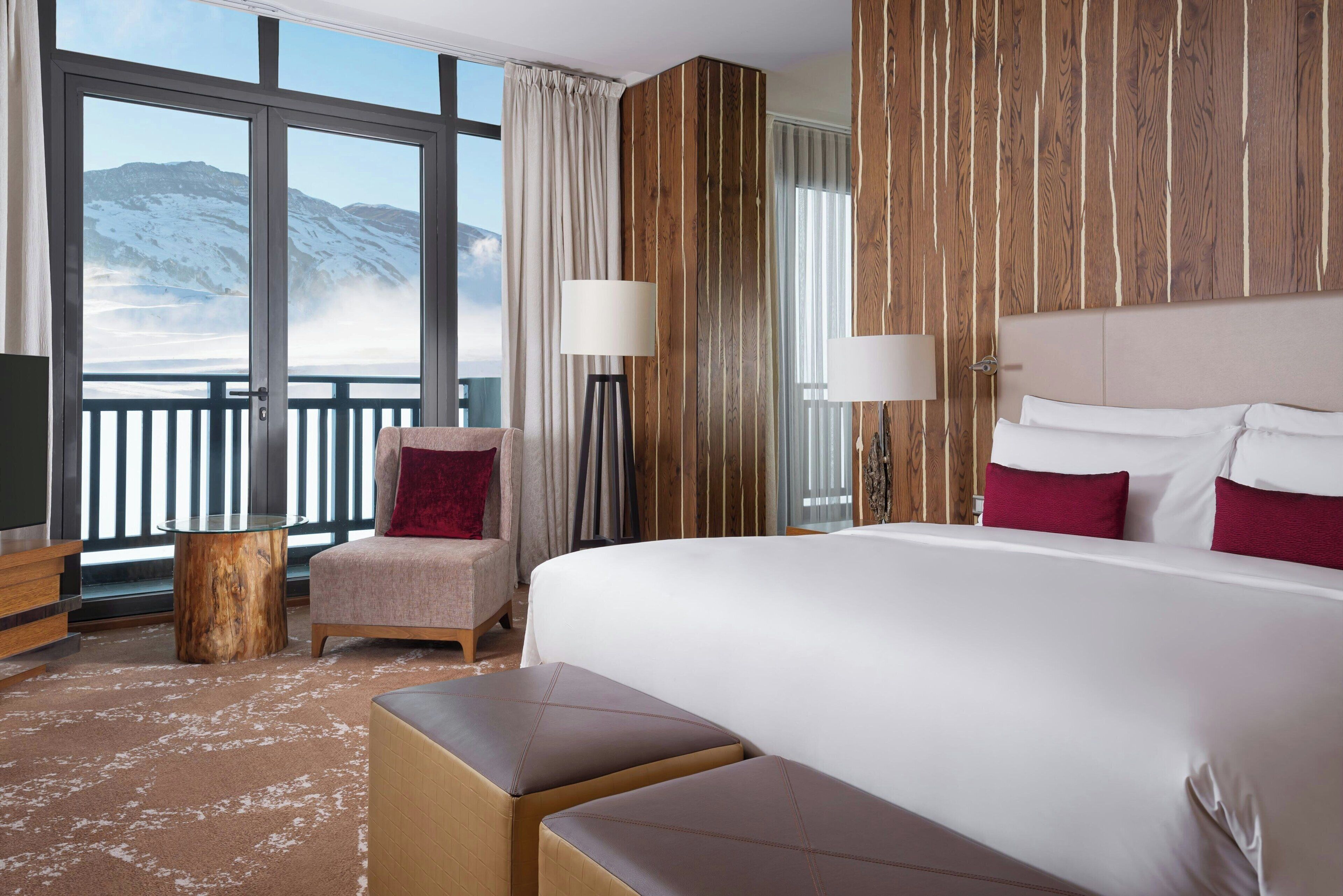 Une chambre d’hôtel avec un grand lit, une chaise, une petite table et une vue sur les montagnes par de grandes fenêtres.