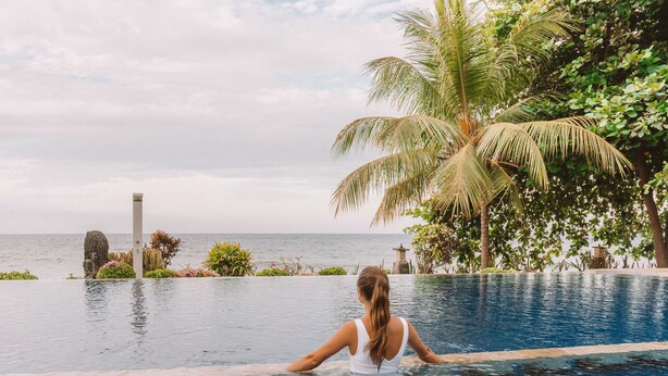 A woman in a white swimsuit sitting at the edge of a pool with a view of the sea and palm trees. - Bondalem Beach Club (Tejakula)