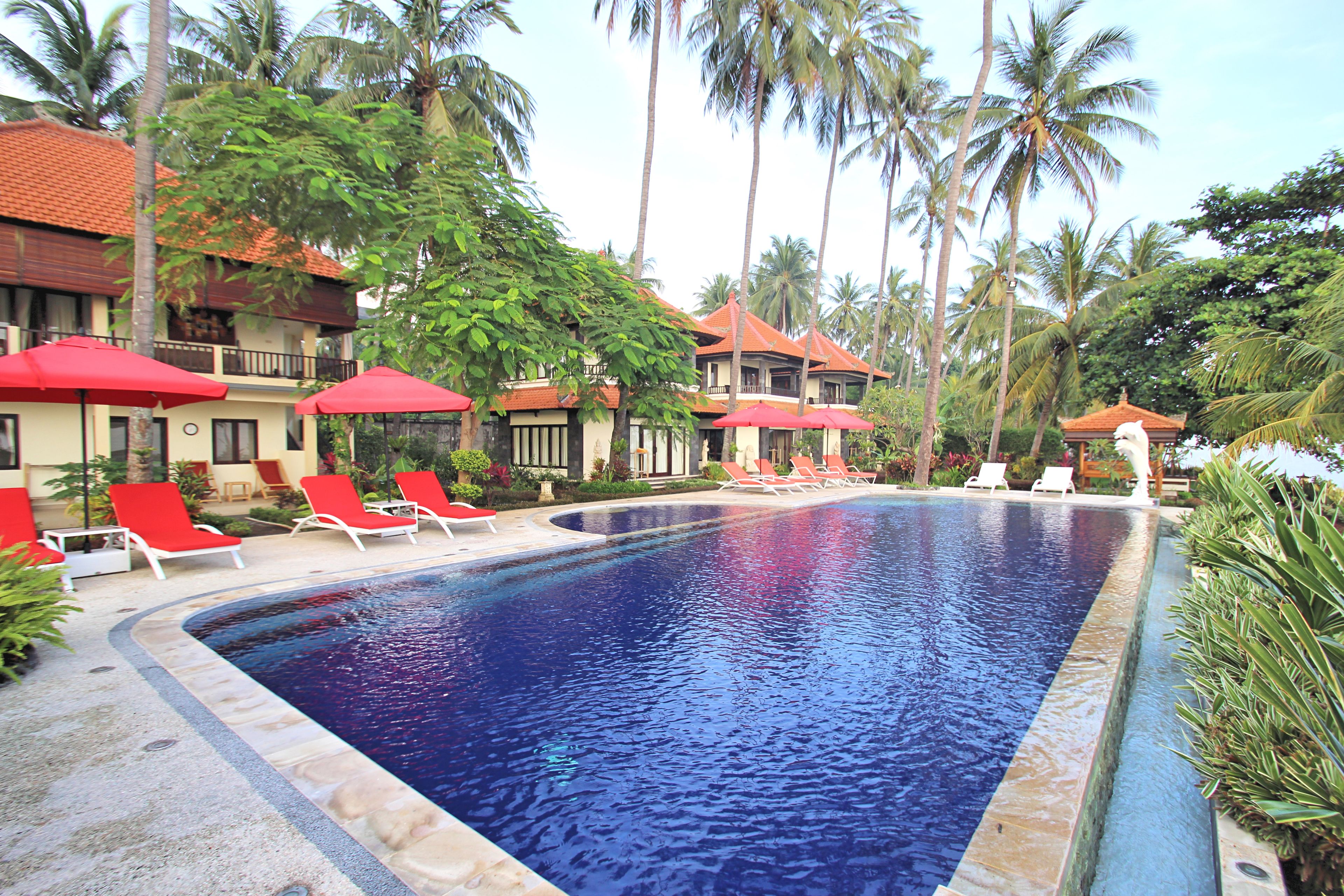 A swimming pool with red umbrellas and lounge chairs, surrounded by palm trees and a building with a red roof.