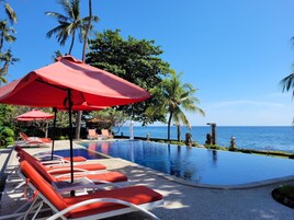 Zona de piscina con sombrillas rojas y sillones de descanso, palmeras y vista al océano.