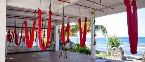 A spacious indoor area with red aerial yoga hammocks suspended from the ceiling, a view of the ocean, and a stone statue in the foreground.
