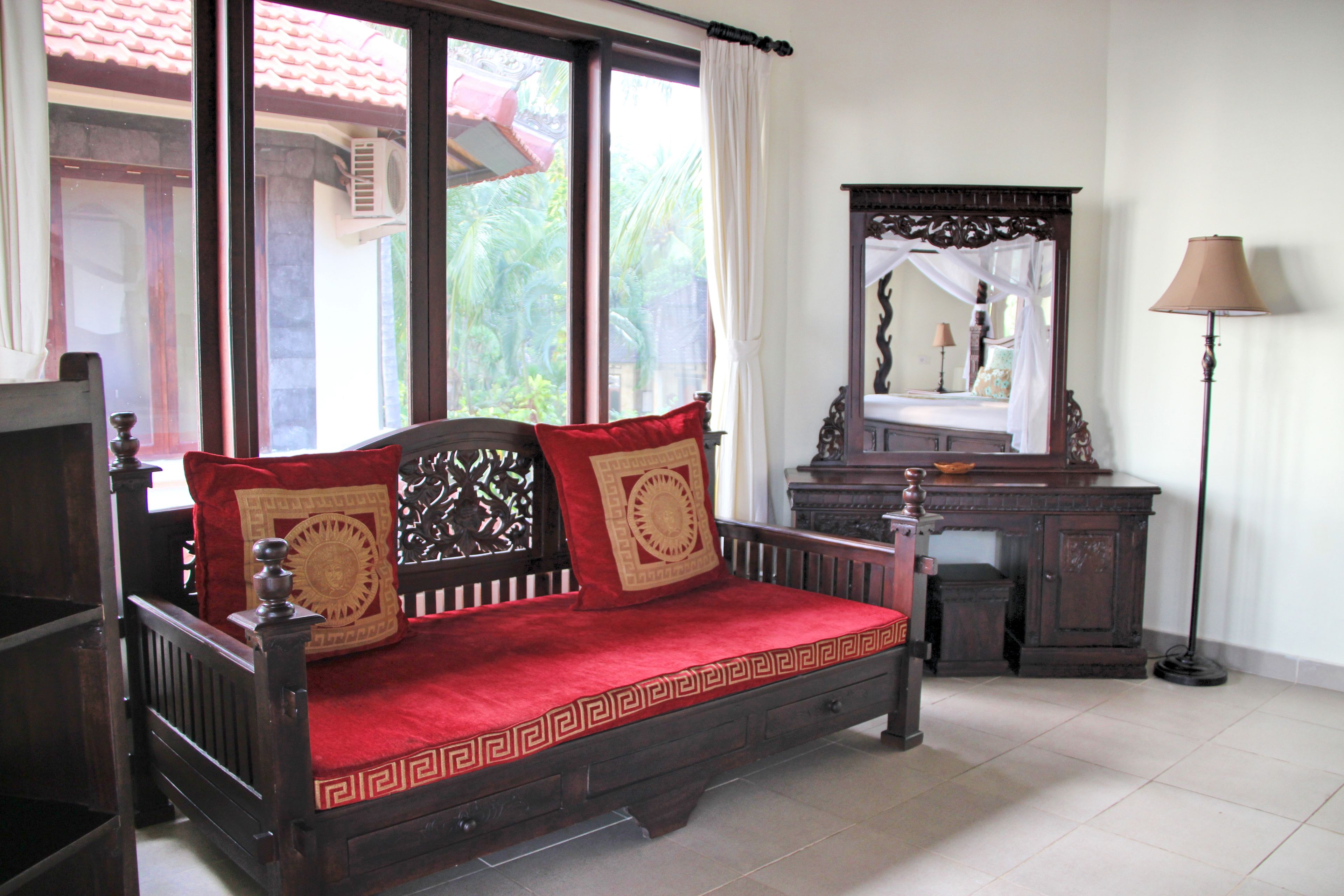 Una cama de madera tradicional con cojines rojos y dorados, una mesita de noche de madera, un espejo y una lámpara.