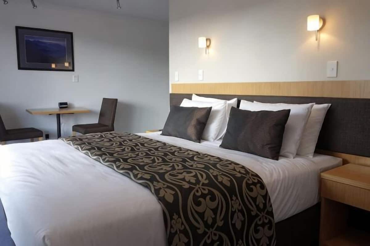 A hotel room with a bed, bedside table, lamp, chair, and a small table with a chair.