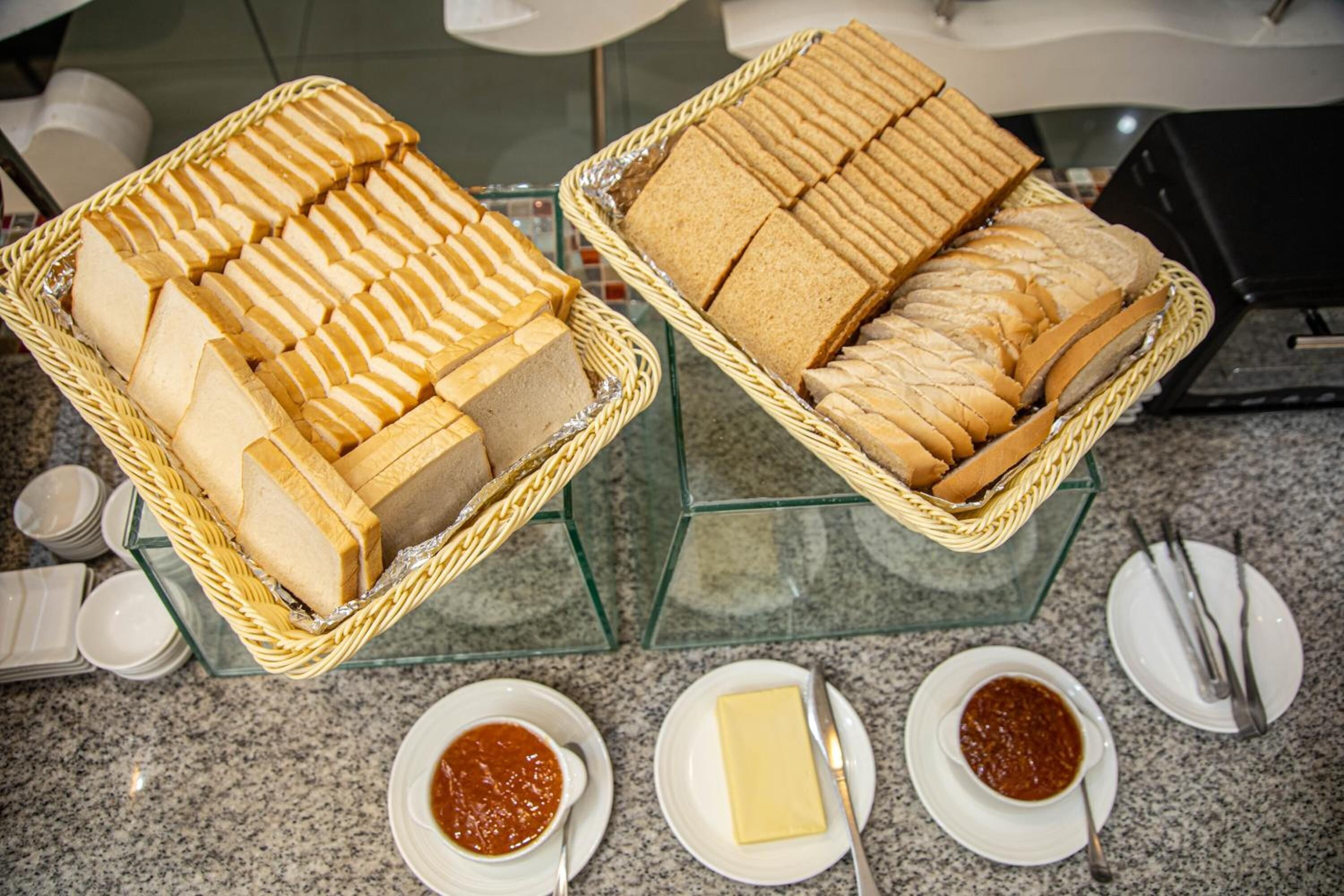 Dos cestas de mimbre repletas de pan en rodajas sobre una mesa de cristal, junto con platos de mantequilla y salsa de tomate.