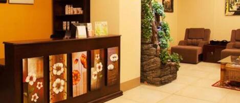 Zona de recepción con mostrador de madera que exhibe arte floral, un pequeño jardín interior y un área de descanso con sofás marrones.