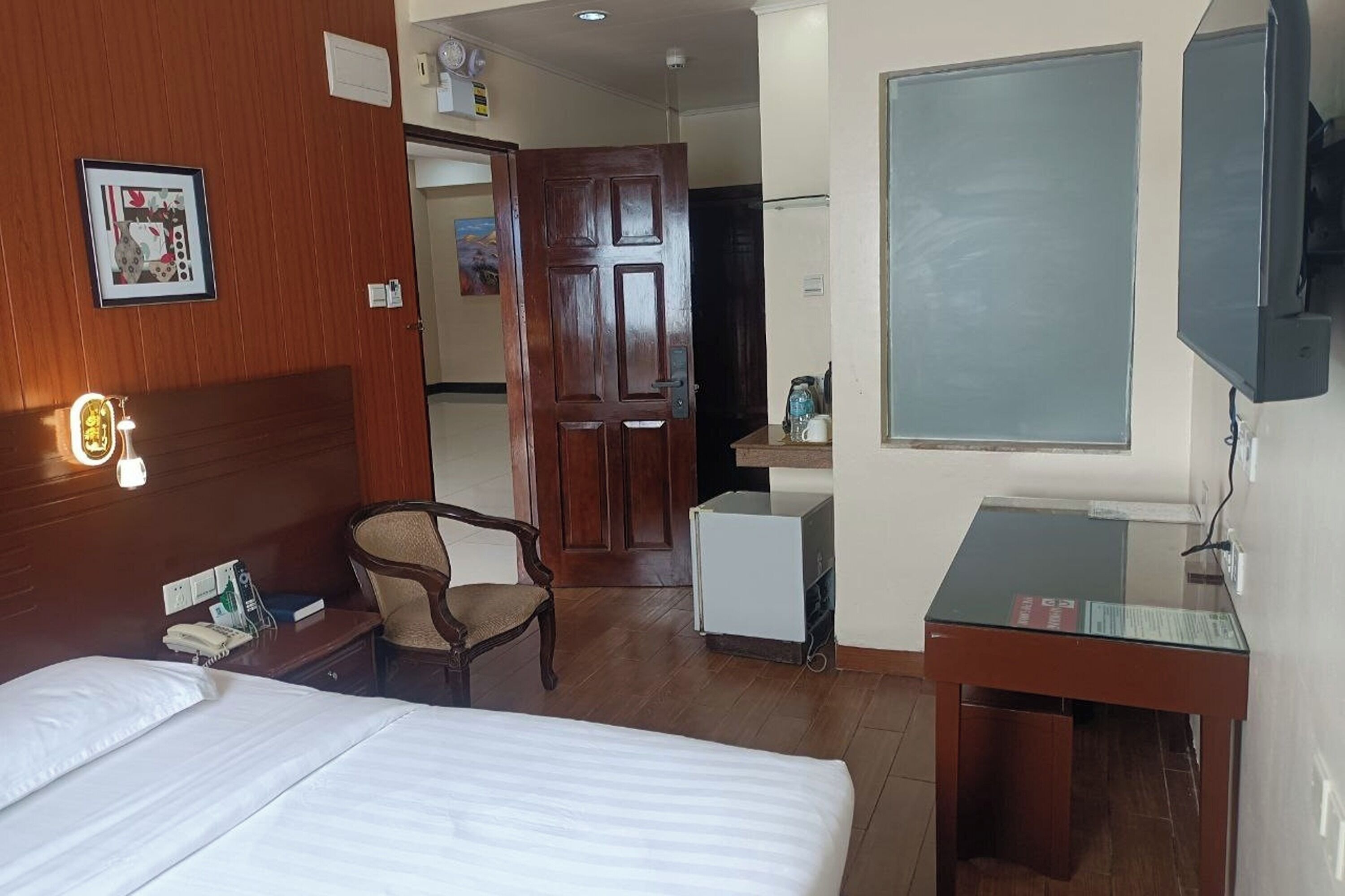 Una habitación de hotel con cama, escritorio, silla, refrigerador, televisor y puerta de madera.