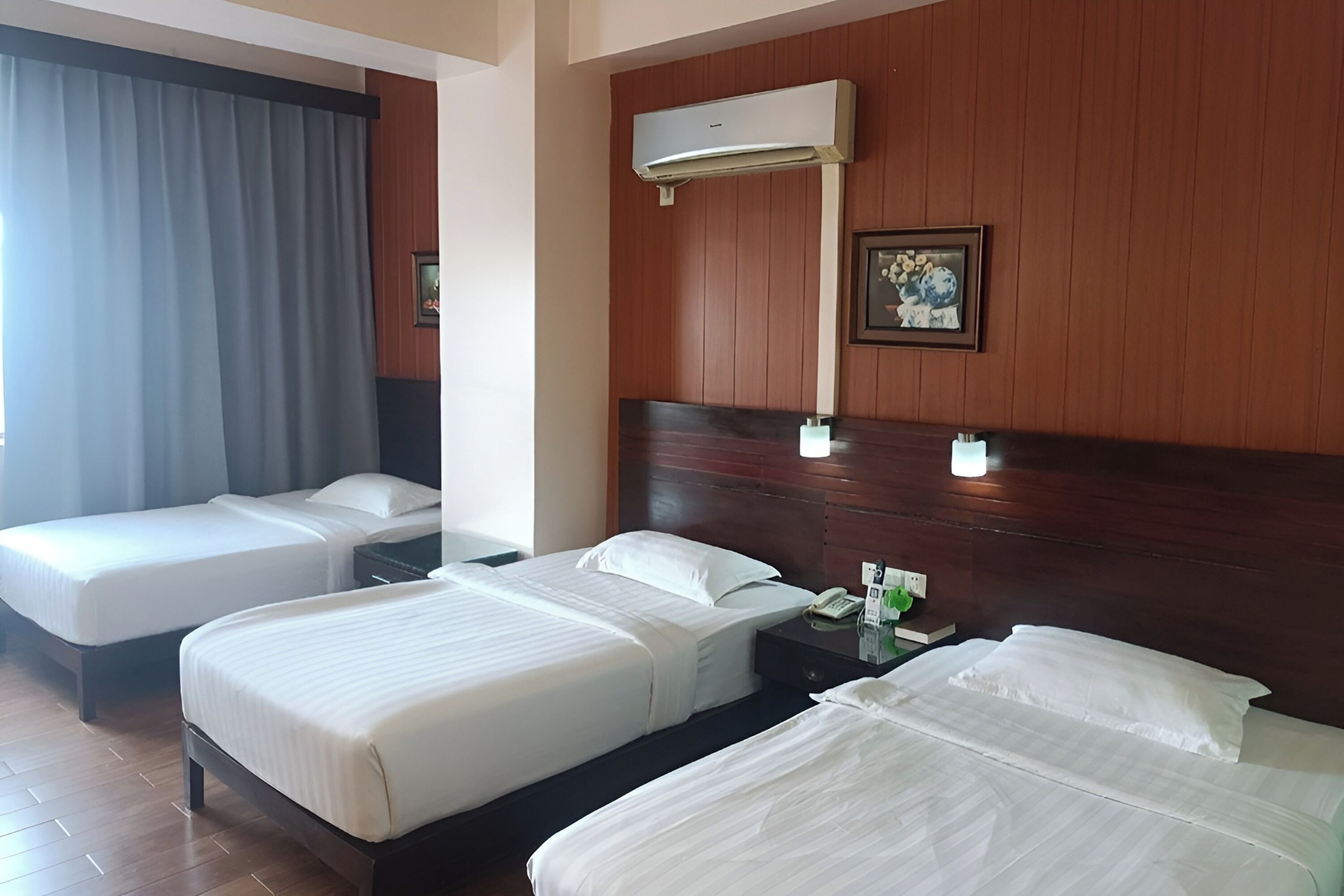 A hotel room with three beds, a wooden headboard, an air conditioning unit, a bedside table with a phone, and a framed picture on the wall.