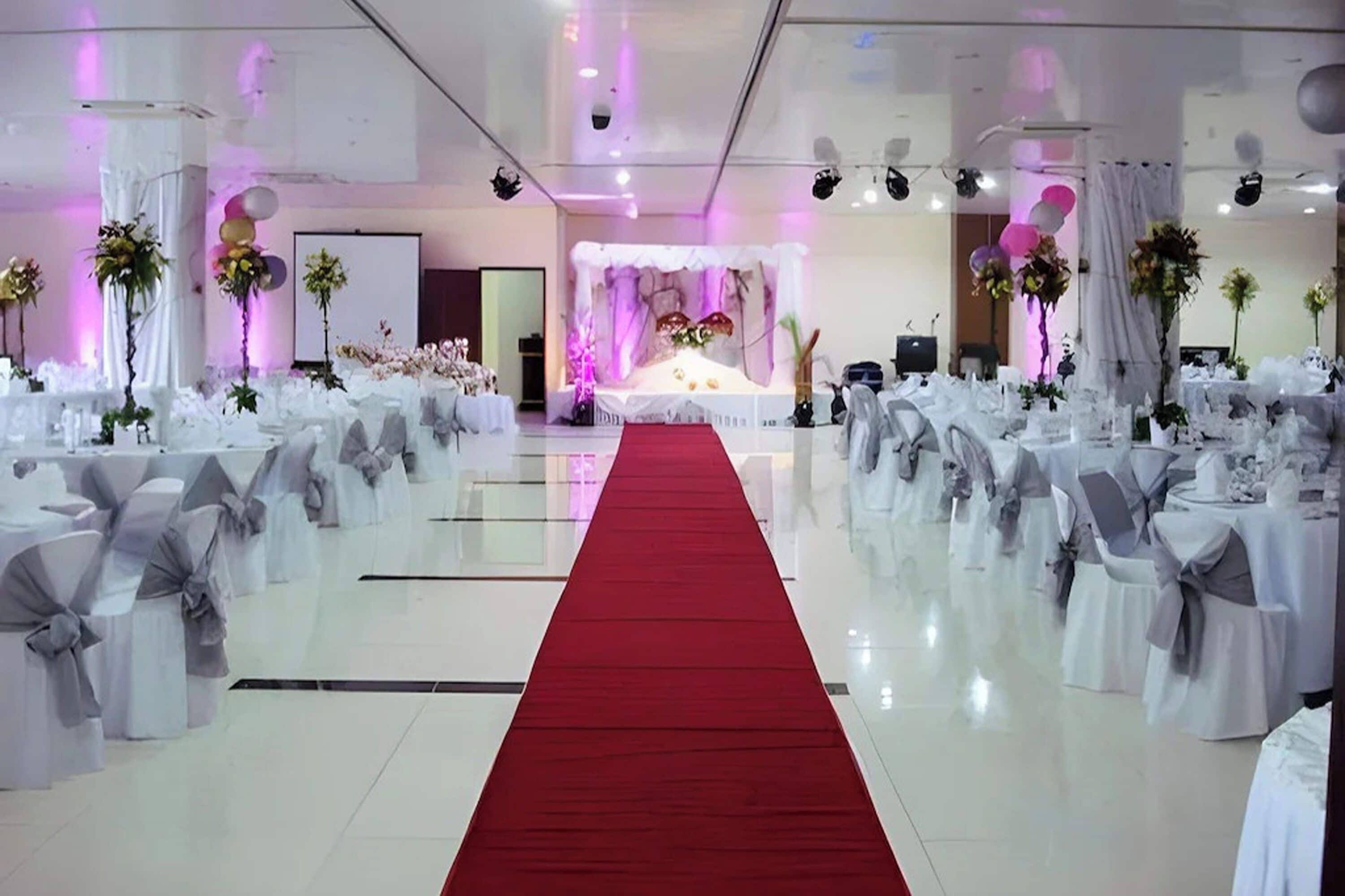 A banquet hall with white tables and chairs, a red carpet, and floral decorations.