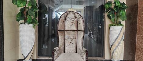 Una silla imponente con respaldo de mármol y asiento mullido, flanqueada por dos grandes macetas con hojas anchas.