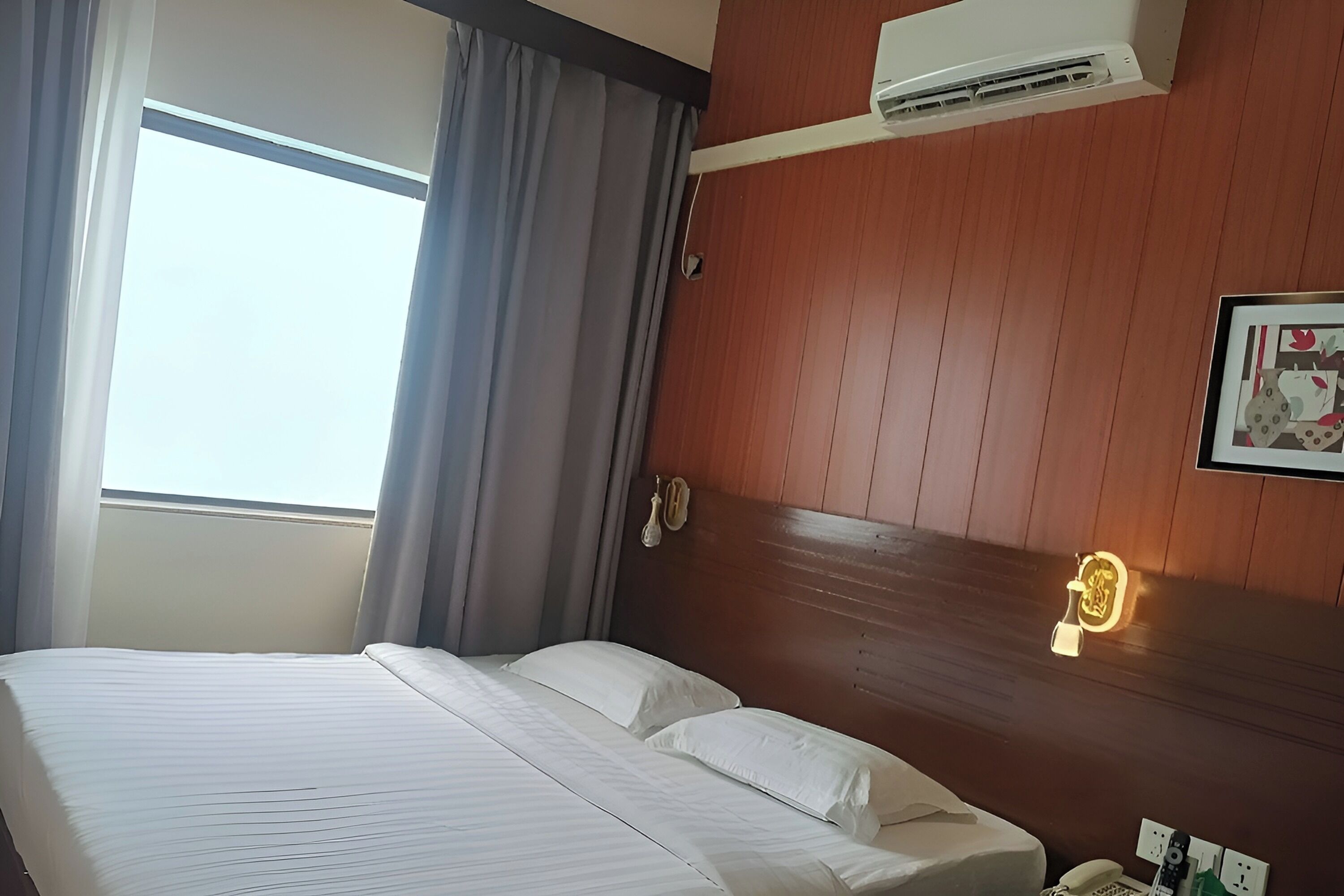 A hotel room with a bed, curtains, an air conditioning unit, a wall-mounted phone, and a light fixture.
