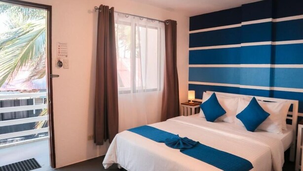 A hotel room with a bed, a nightstand, a window with curtains, and a view of palm trees outside. - 8Colors Beach House (Boracay Island)