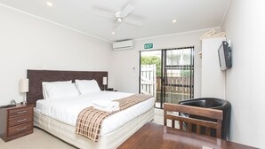 A hotel room with a bed, bedside table, lamp, ceiling fan, air conditioning unit, and a view of the outside.