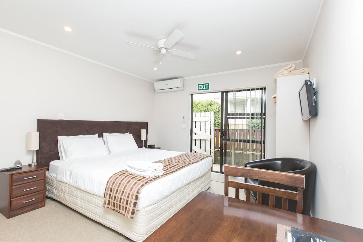 Habitación de hotel con cama, mesita de noche, lámpara, ventilador de techo, aire acondicionado y vistas al exterior.
