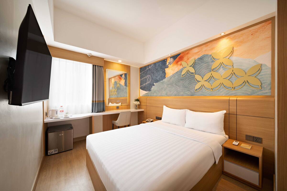 A hotel room with a large bed, a desk with a chair, a television, and a wall with a floral artwork.