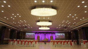 Un salón de banquetes con filas de sillas blancas y cintas rojas, un escenario con un telón de fondo morado y varios candelabros.