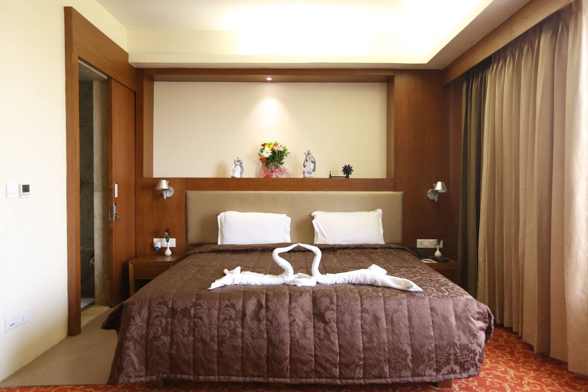 A hotel room with a large bed, two white pillows, a wooden headboard, a nightstand with a lamp, and a small table with a vase of flowers.