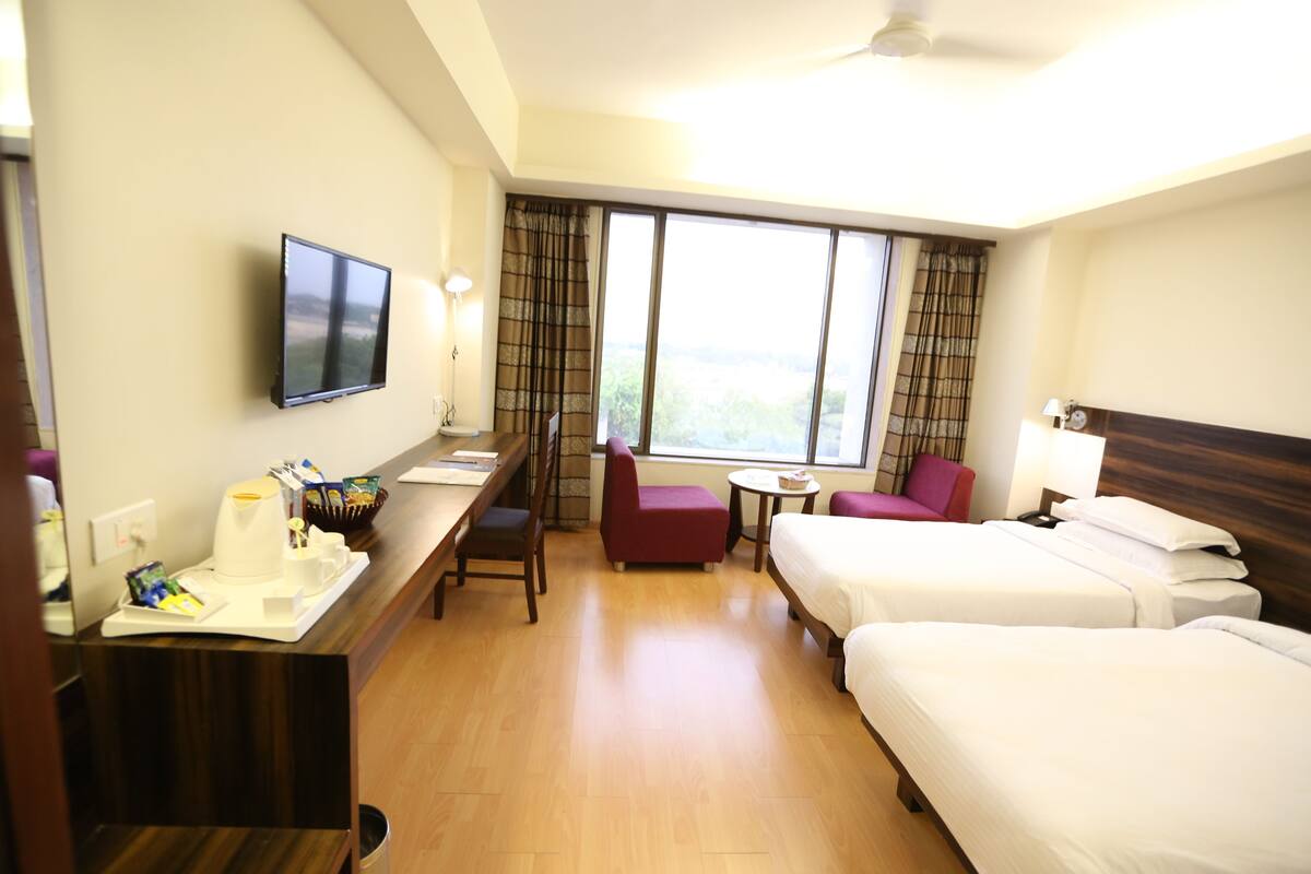 A hotel room with two beds, a desk with a coffee maker, a TV, and a view of the outdoors.