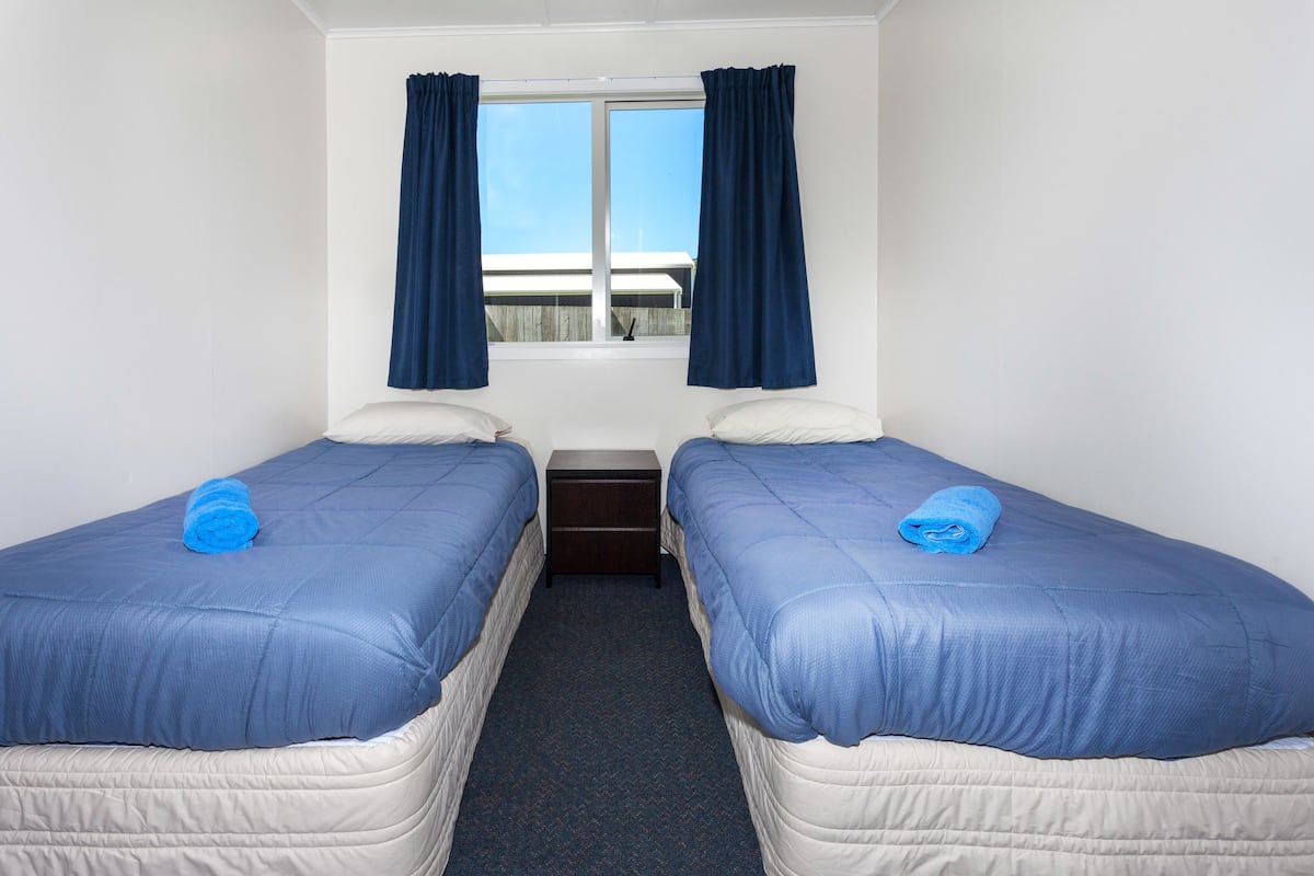 Une chambre avec deux lits, chacun recouvert d’une couverture bleue et doté d’un oreiller blanc. Une petite table de chevet se trouve entre les lits, et il y a une fenêtre avec des rideaux bleus.
